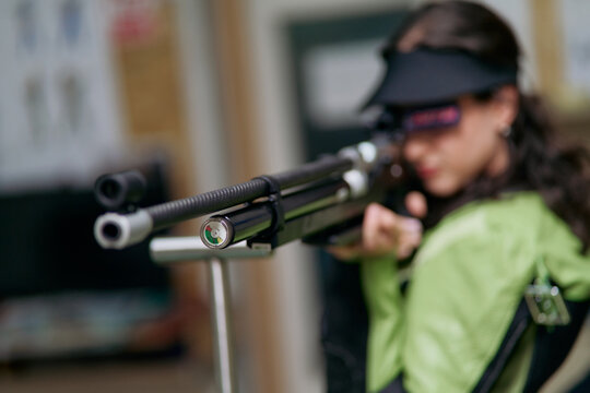 Girl aiming air rifle during shooting training, focus on the rifle, practice for sport competition, precision and focus, indoor shooting range, blurred background.