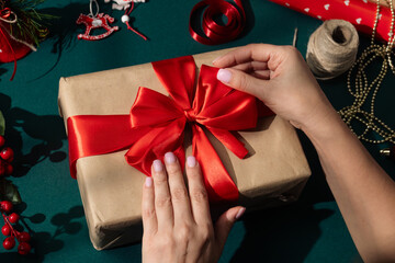 Female hands adjusting a red satin ribbon bow on a kraft paper wrapped Christmas present surrounded by festive decorations, and wrapping materials on a green background.