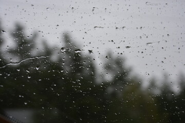 Raindrops on a window with a blurred, dark forest and a grey, overcast sky in the background. A moody, melancholic, and atmospheric scene