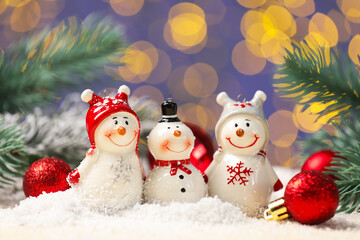 Cute decorative snowmen, fir tree branches and red baubles on artificial snow against blurred lights, closeup