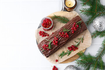 Delicious Yule log (traditional Christmas cake) with redcurrant, candle and fir tree branches on white tiled table, flat lay. Space for text