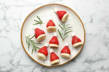 Christmas food. Santa hats made of marshmallows, strawberries, bananas and rosemary on white marble table, top view