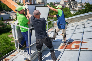 Workers lifting up photovoltaic solar panel on metal rooftop of house with assistance of crane...