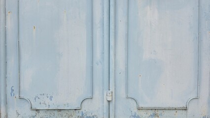 Old light blue metal door with peeling paint, rust stains, and embossed panel details, showing aged texture and natural weathering marks on the surface.