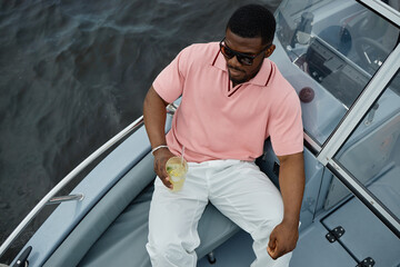 Young adult Black man sitting on boat holding drink with lemon slice, wearing sunglasses, relaxing by water, looking down, enjoying leisure time during summer outing