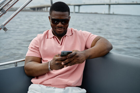 Black young adult man sitting on boat using smartphone, wearing sunglasses, smiling while looking at device, water and bridge visible in background, relaxed outdoor setting