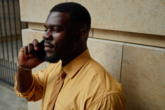 Young adult Black man standing outdoors talking on smartphone, wearing formal shirt and tie, looking away from camera with slight smile, urban background visible