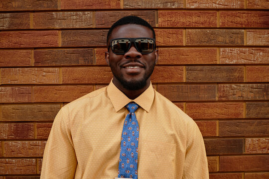 Portrait of young adult Black man smiling and wearing sunglasses while standing against brick wall, facial hair visible, looking directly at camera, formal attire evident