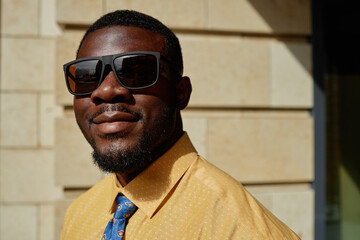 Portrait of young adult Black man wearing sunglasses smiling confidently outdoors, short beard visible, standing against stone wall, sunlight highlighting facial features