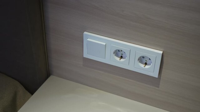 Light switch and sockets on wooden wall near bed headboard closeup. Electrical equipment plugs and control key in modern hotel bedroom