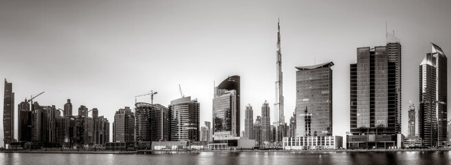 Naklejka na ściany i meble Monochrome city skyline with tall skyscrapers reflecting on calm water under a cloudy sky, Dubai Marina bay UAE Naklejka na ściany i meble Monochrome city skyline with tall skyscrapers reflecting on calm water under a cloudy sky, Dubai Marina bay UAE