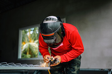 Welder wearing a red long-sleeved shirt and a protective mask is welding a steel grating, Industrial Welder With Torch