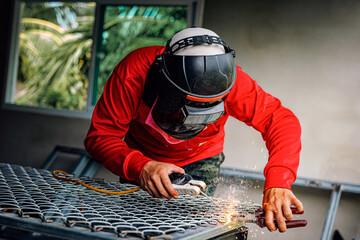 Welder wearing a red long-sleeved shirt and a protective mask is welding a steel grating, Industrial Welder With Torch