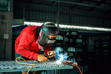 Welder wearing a red long-sleeved shirt and a protective mask is welding a steel grating, Industrial Welder With Torch