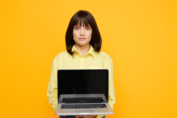 Fototapeta premium A calm woman stands against a bright yellow backdrop, holding an open laptop toward the camera. The image conveys focus, remote work, and digital creativity.