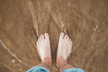 Caucasian male bare feet and pulled up jeans, standing on a sandy beach with shallow tidal waves sea water reaching the shore. Top down, close up view