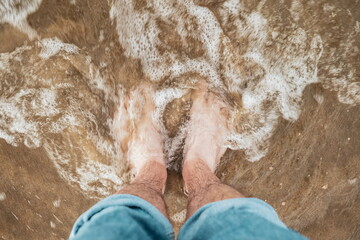 Caucasian male bare feet and pulled up jeans, standing on a sandy beach with shallow tidal waves sea water reaching the shore. Top down, close up view