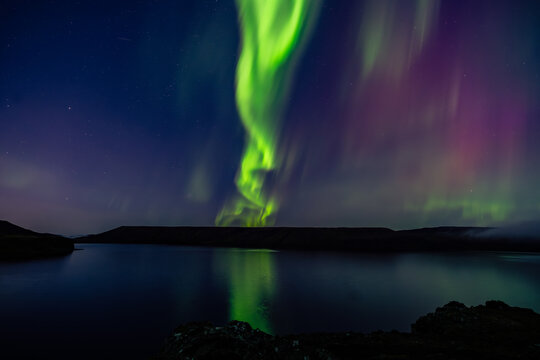 View of ethereal green and purple Northern Lights dancing above the still, reflective waters under a starlit sky, Northern Lights, Grindav&iacute;kurbaer, Iceland.