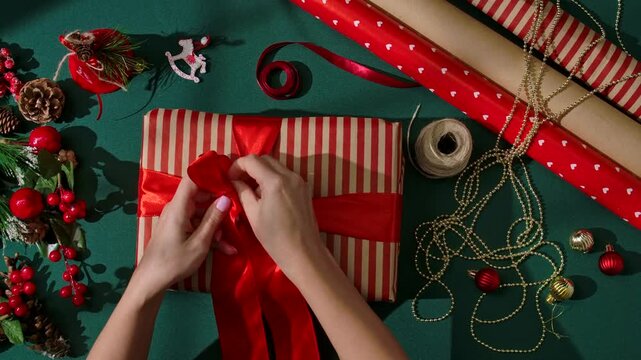 Top view of a woman hands tying a red satin ribbon on a striped gift box among scissors, twine, and decorations on a green background for Christmas crafts.