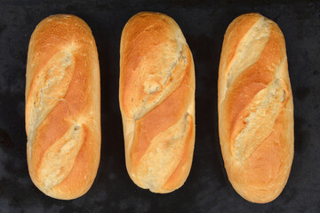 An overhead shot of three freshly baked, golden-brown bread rolls lying side-by-side on a dark, slightly textured baking surface