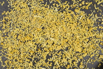 A horizontal, full-frame macro shot of finely curled, dried yellow lemon zest scattered across a dark surface, creating a rough texture