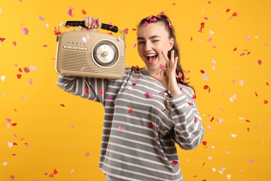 Excited young woman with radio receiver showing ok gesture among falling confetti on yellow background
