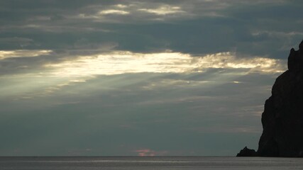Godrays silhouette rockformation over the dark ocean at dramatic sunset with powerful sunbeams piercing the heavy storm clouds - Powered by Adobe