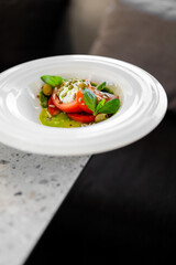 Fresh caprese salad with sliced tomatoes, creamy burrata cheese, pesto, basil leaves, and seeds, served on a white plate on a dark marble table
