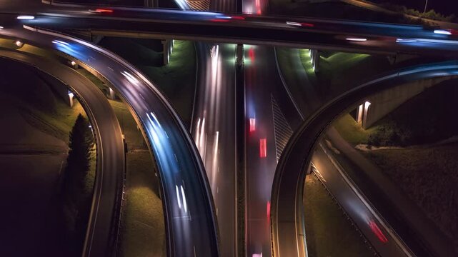 Bright lights and flowing cars fill a busy highway interchange at night. The intricate layout and reflections create a vibrant urban scene, showcasing the motion of vehicles