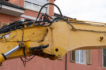 Yellow excavator joint with hydraulic tubes, where hydrauilic fluid is coming out and spreading over the arm. Old reddish house in the background.
