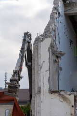 Grab of excavator is holding a reddish corrugated steel sheet, close to the wall of a tear down building. Cloudy sky. Vertical shot.
