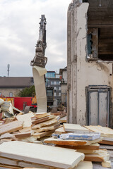 Grab of excavator is holding a white steel sheet, close to the wall of a tear down building. Cloudy sky. Vertical shot. Rubble and debris in the foreground.