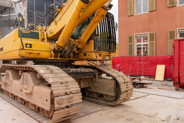 Close up on huge yellow excavator with chains and operator protection on a construction site. Tear down building and building still in use in the background.