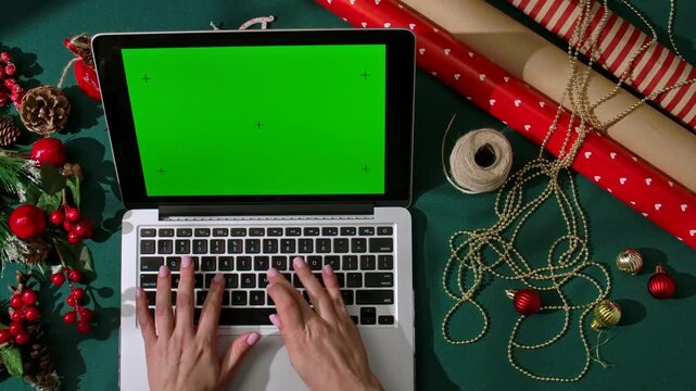 Top view of hands typing on a laptop with a chromatic green screen on a dark green festive background surrounded by packaging materials and decorations. The perfect layout for holiday shopping or