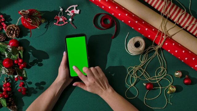 Top view of women hands swiping a smartphone with a green screen on a dark green festive background. The phone is surrounded by festive packaging materials, and Christmas tree decorations.