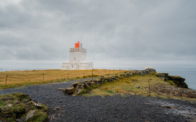 View of a striking white lighthouse with a vibrant orange top stands proudly against the moody sky in Reynisfjara Beach, Myrdalshreppur, Iceland.
