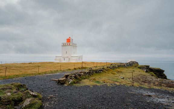 View of a striking white lighthouse with a vibrant orange top stands proudly against the moody sky in Reynisfjara Beach, Myrdalshreppur, Iceland.