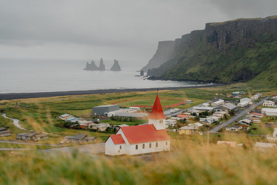Fototapeta View of a quaint white church with a brilliant red roof nestled amidst a village by a black sand beach and jagged cliffs, Reynisfjara Beach, Myrdalshreppur, Iceland.