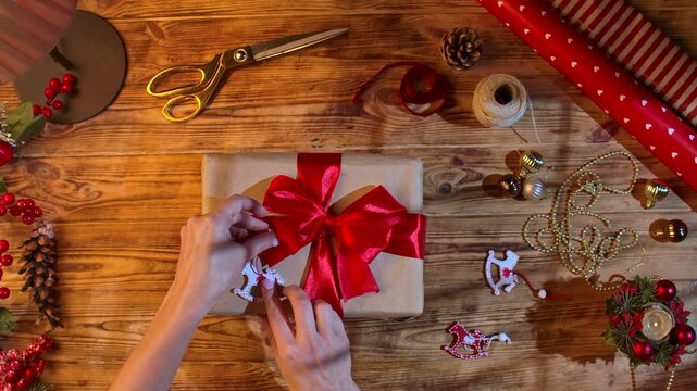 Top view of a woman hands decorated with a toy wooden horse on a gift box, among decorations on a rustic wooden table for Christmas crafts.