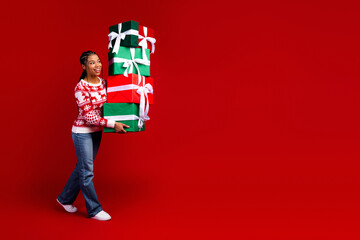 Funny holiday moment a young woman in a festive sweater carries a stack of bright presents against...