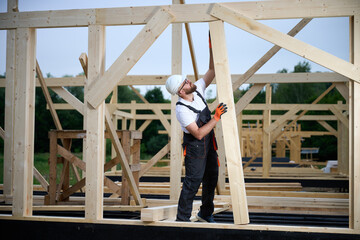 Construction worker installing wooden beam at house frame construction site. Carpenter in safety gear building timber structure for new home outdoors.