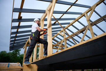 Construction worker using an electric screwdriver while building a wooden frame structure. Man in protective helmet, gloves. Concept of construction, carpentry, engineering and building process.