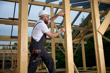 Construction worker measuring wooden beam with tape measure at building frame. Builder in safety...