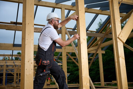 Construction worker measuring wooden beam with tape measure at building frame. Builder in safety helmet working on wooden house construction site outdoors.