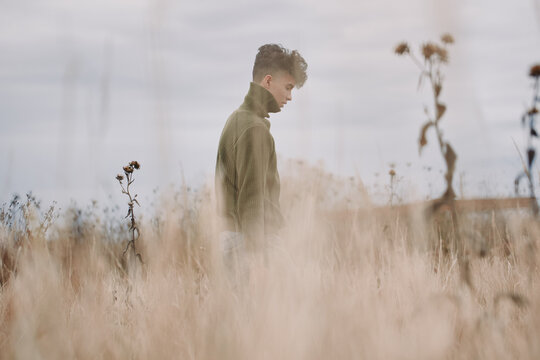 A young man in a knit sweater walks through tall dry grass in a muted field, photographed with natural light to convey authenticity and credibility in a simple, candid portrait of solitude.