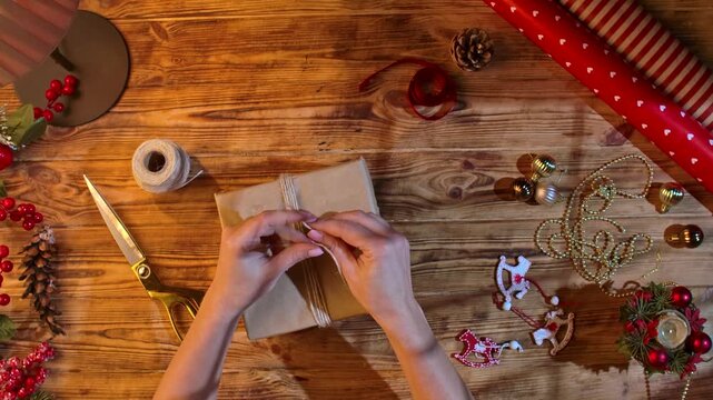 Top view of a woman hands tying twine around a Christmas gift in craft packaging on a wooden table with a red ribbon, pine cone, and gold scissors. A cozy moment of wrapping gifts yourself.