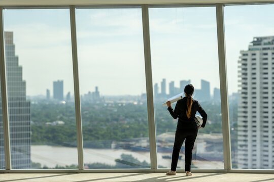 architect woman in suit thinking planning thoughtful inspection on empty office interior near glass window. office interior standing thinking planning thoughtful with blueprint on skyscraper floor