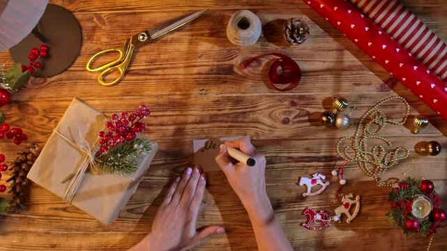 Top view of hands writing Merry Christmas on a kraft paper gift tag next to a wrapped gift, ribbons, and decorations on a wooden table. Cozy holiday packaging and a handmade greeting concept.