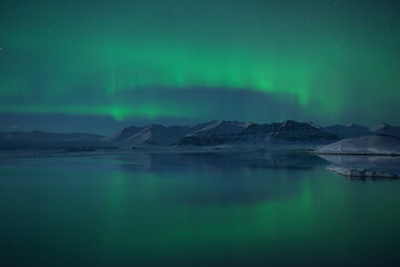 View of the ethereal green aurora borealis dances above the icy lagoon, reflecting in the still waters beneath the snow-capped mountains, Jokulsarlon, Sveitarfélagio Hornafjorour, Iceland.
