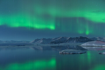 View of emerald aurora borealis dances above the glacial lagoon, reflecting in the still, dark waters amidst snow-capped mountains, Jokulsarlon, Sveitarfélagio Hornafjorour, Iceland.